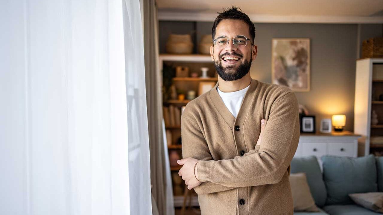 Junger Mann in einer Strickjacke steht in einem Wohnzimmer. Er lacht zufrieden in die Kamera.