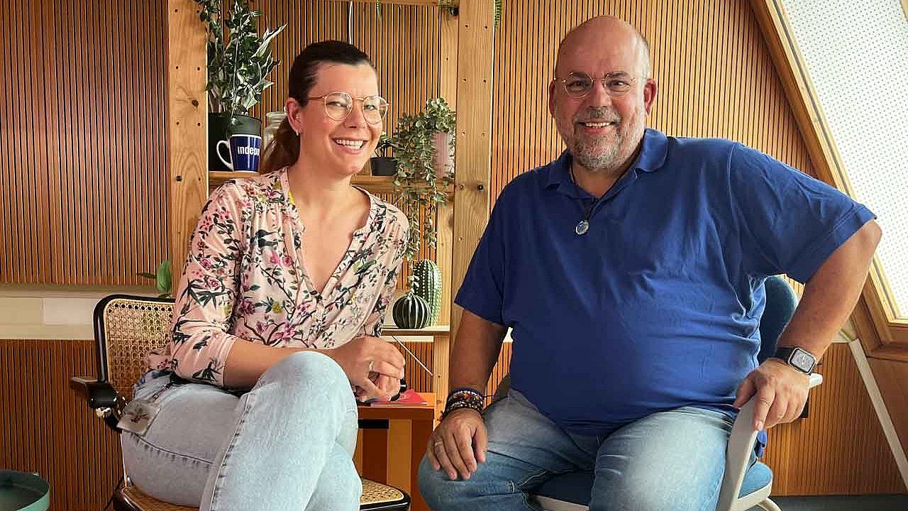 Host Charlotte Mattes (links), rechts Pfarrer Nulf Schade James, auf Stühlen, im Podcaststudio in Frankfurt Heddernheim. Beide lächeln.