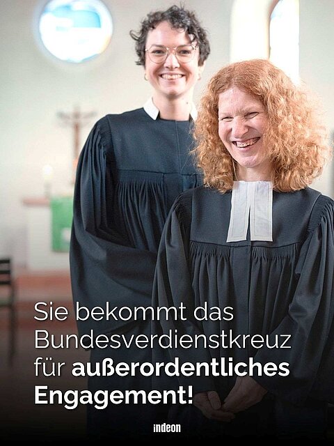 Zwei Pfarrpersonen im schwarzen Talar in einem Kirchengebäude: im Hintergrund eine Person mit kurzen dunklen Locken, im Vordergrund Brigitte Buchsein, mit roten lockigen Haaren, herzlich lächelnd. Text: Sie bekommt das Bundesverdienstkreuz für außerordentliches Engagement.