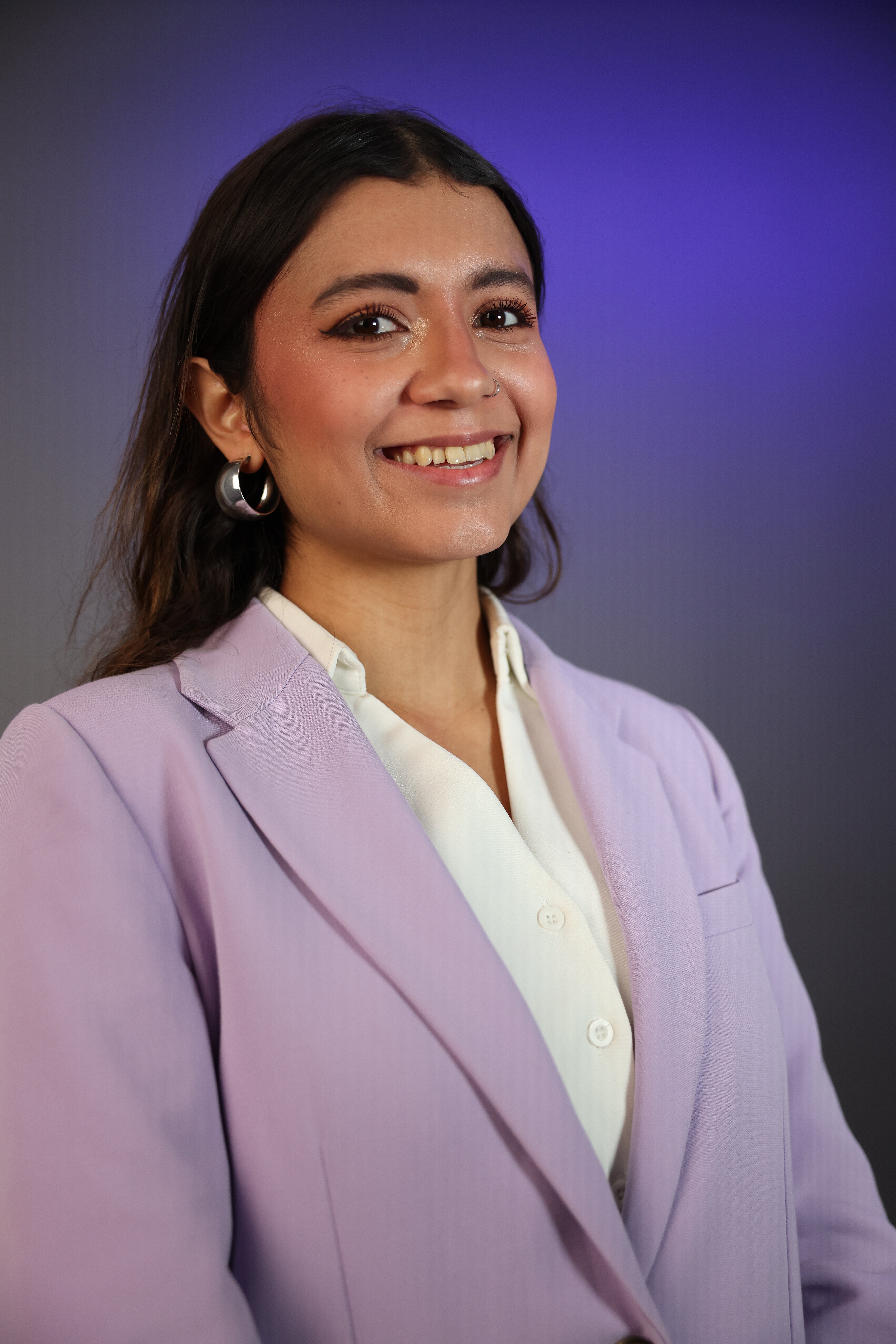 Isabella, steht in einen Fliederfarbenen Blazer und weißer Bluse vor Violettem Hintergrund und und lächelt in die Kamera. 