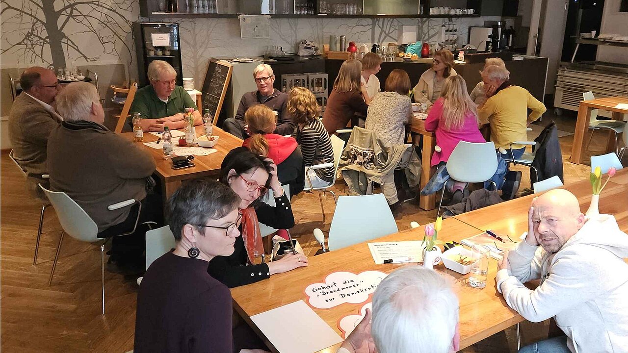 Kleine Gruppen von Menschen sitzen an verschiedenen Tischen. Sie sind ins Gespräch vertieft. Auf den Tischen Getränke, Knabbereien, Blumen und beschriftete Plakate, die das Thema der Diskussion vorgeben, z.B.: Gehört die Brandmauer zur Demokratie. Im Hintergrund ist eine graue Barzeile zu erkennen.