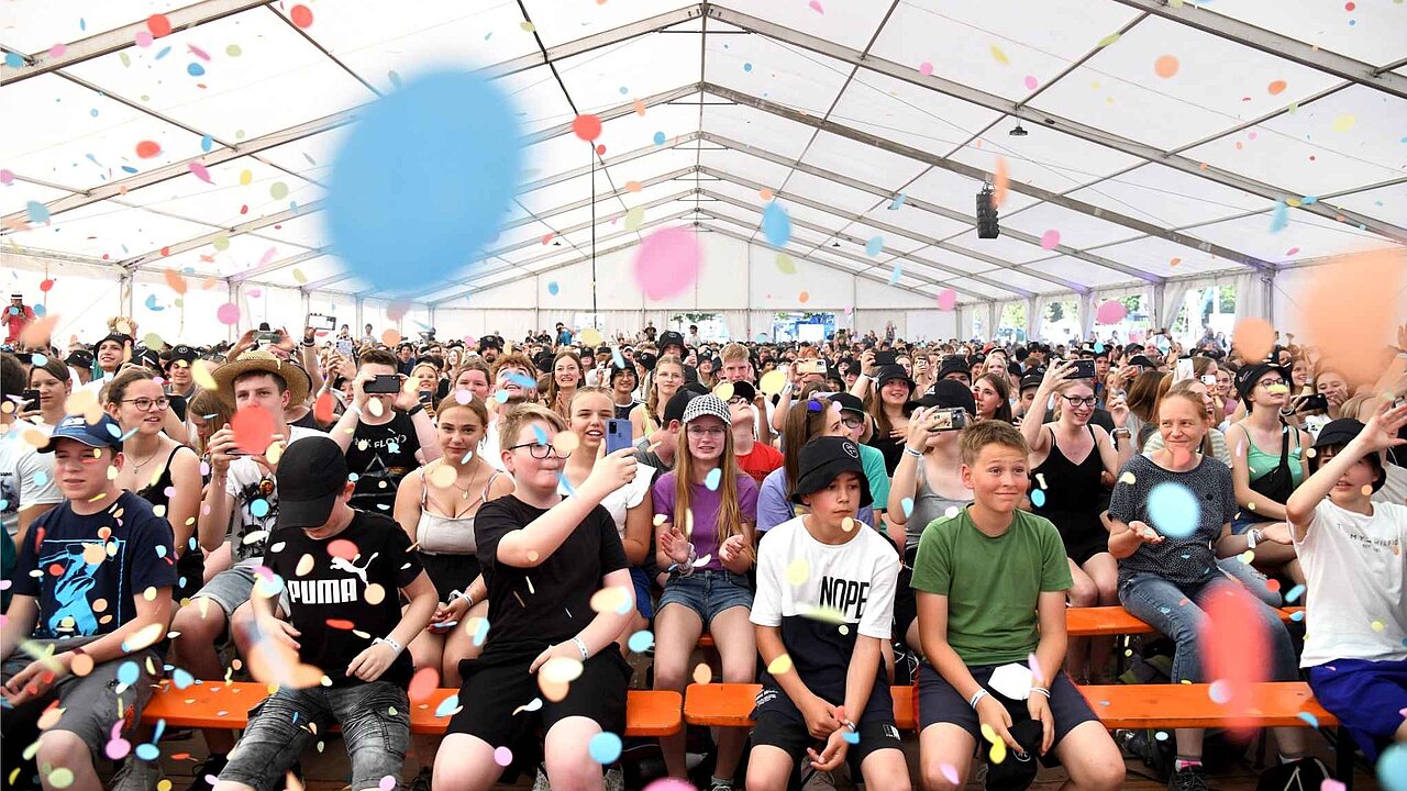 Eine Menschenmenge mit vielen Kindern und Jugendlichen in einem Festzelt. In der Luft sind viele Luftballons
