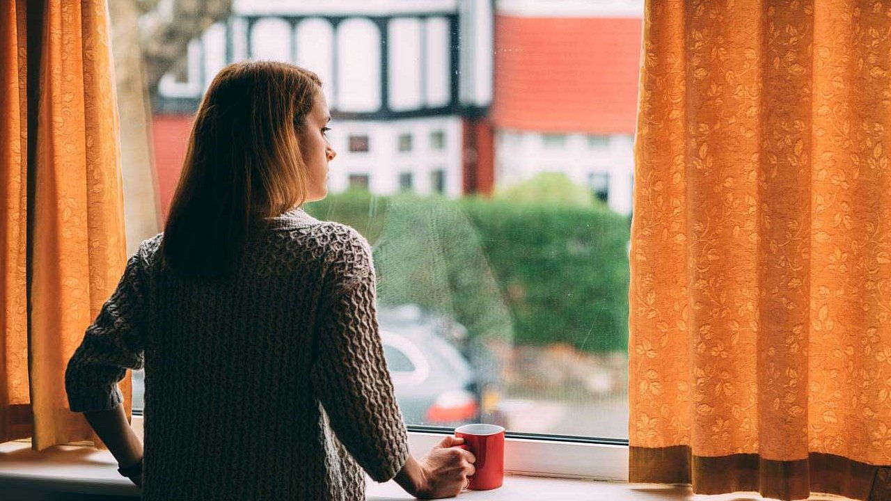 Eine Frau steht am Fenster ihrer Wohnung und schaut nach draußen. Die Kontaktbeschränkungen durch Corona haben viele von uns unter Stress gesetzt.