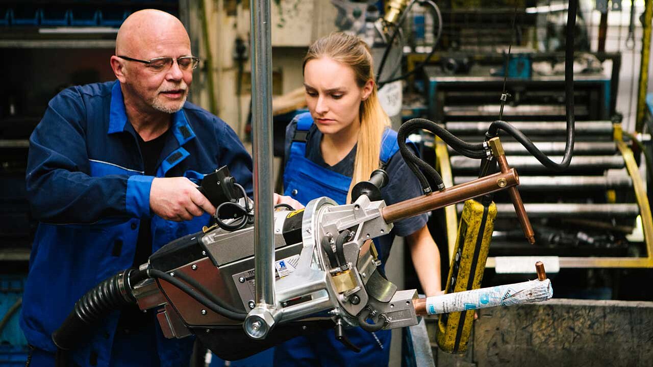 Ein erfahrener und älterer männlicher Mitarbeiter gibt der weiblichen Auszubildenden Anweisungen in der Metallindustrie