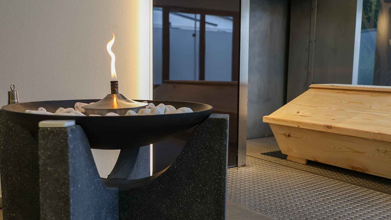 Sarg vor der Einäscherungsanlage, links daneben Brennt eine Flamme, die Öffnung der Anlage ist mit Ornamenten verziert 