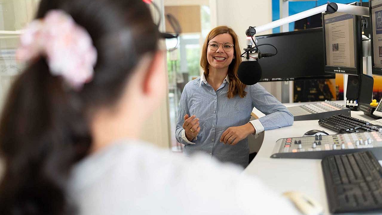 Im Vordergrund, unscharfer Pferdeschwanz, mit rosa Haargummi, im Hintergrund Host Charlotte Mattes, lächelt und gestikuliert im Aufnahmestudio.