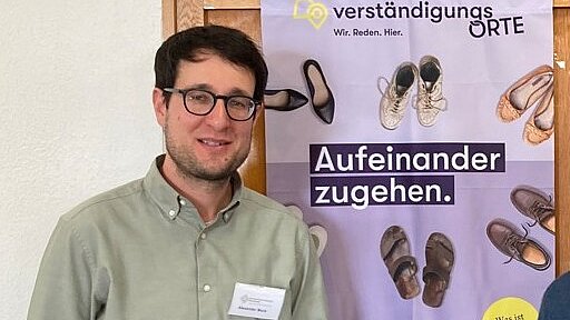 Ein Mann mit grünem Hemd, dunkler Brille und dunklen Haaren steht vor einem Poster zu Verstädnigungsorten