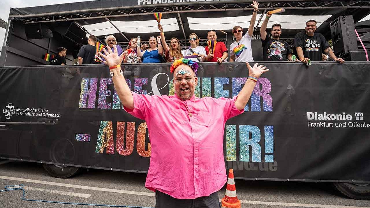 Nulf Schade-James, Pfarrer seit 1989 in der Kirchengemeinde Frieden und Versöhnung in Frankfurt, steht zu Beginn der Christopher Street Day Parade (CSD) in Frankfurt vor dem LKW der evangelischen Kirche. Das Motto des CSD, „Here and queer - auch wir“, steht auf den großen Schildern an den Längsseiten des Lkw, auf dem Pfarrpersonen, Gemeindemitglieder und Kolleg*innen anderer Berufe mitfahren (Foto vom 15.07.2023).
