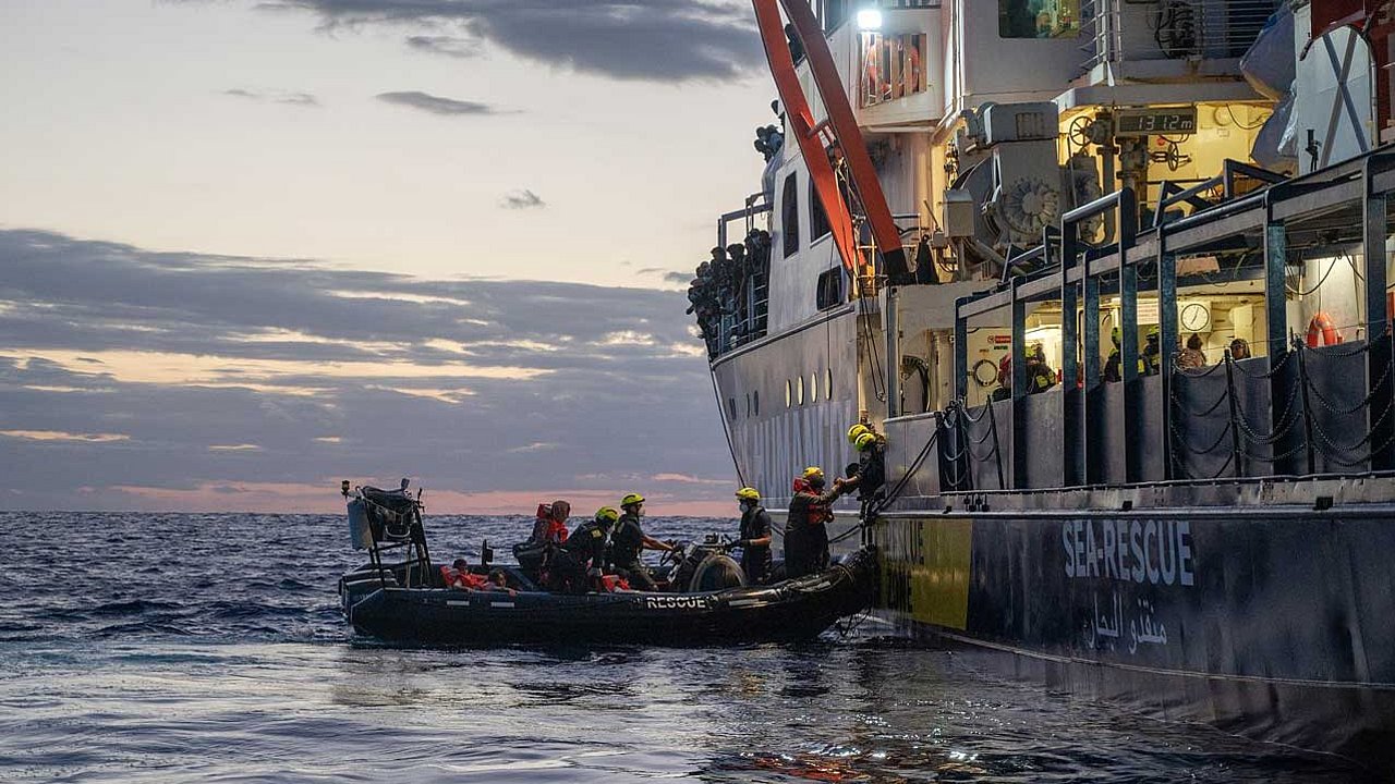 Das Rettungsschiff von der Seite und ein angelegtes Beiboot, von dem Menschen auf das Schiff umsteigen.