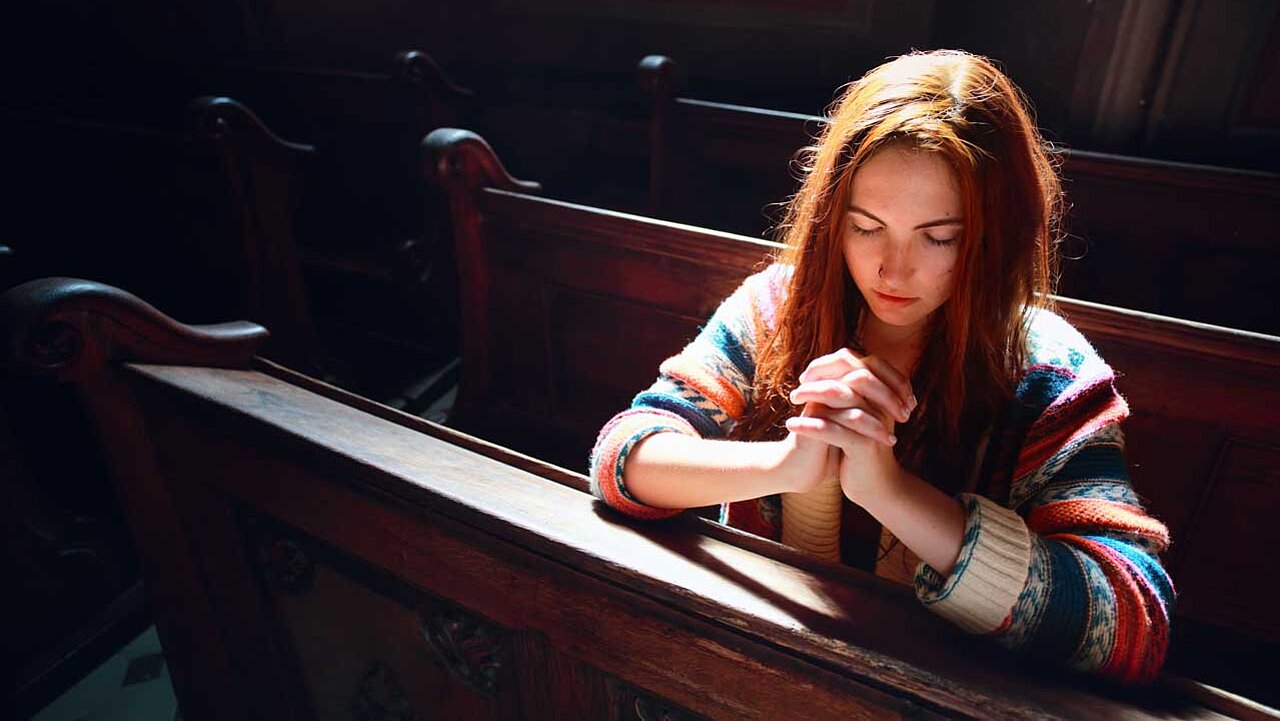 Junge Frau alleine in einer Kirchbank ins Gebet versunken. Von hinten fällt Licht auf ihr Haar.