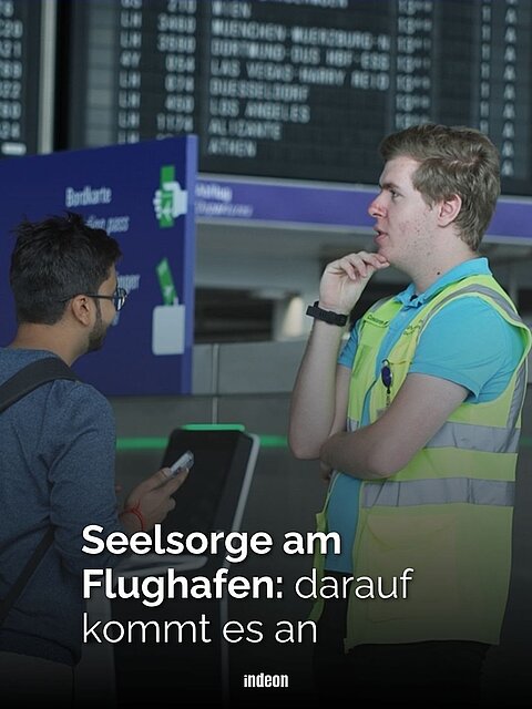 Zwei Männer unterhalten sich, links ein Reisender am Flughafen mit blauem Pulli, Rucksack, Brille und schwarzem Bart und Haaren. Rechts Seelsorger Constantin, groß, dunkelblond, mit hellblauen Poloshirt und gelber Warnweste. Im Hintergrund die Flugtafel mit Starts und Landungen. Text: Seelsorge am Flughafen, darauf kommt es an.