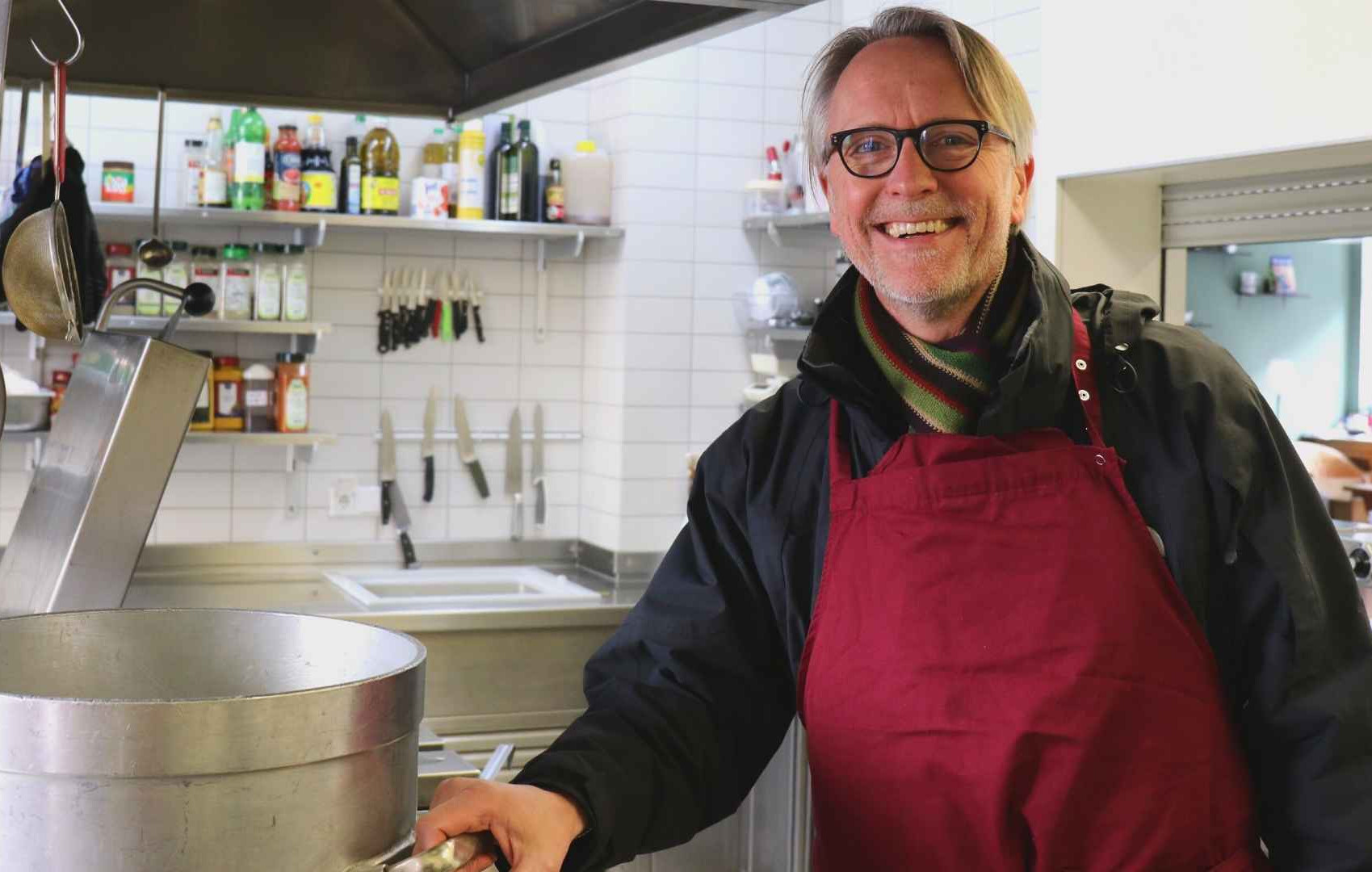 In der Küche der ada_kantine steht Thilo Schwarmann, ein Mitarbeiter, neben einem großen dampfenden Kochtopf. Im Hintergrund sind ein paar Küchenutensilien, unter anderem Messer, aber auch Gewürze.