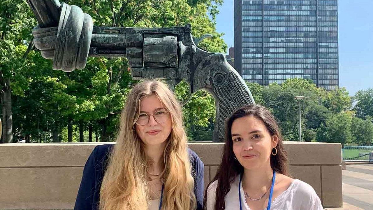 die beiden Studentinnen stehen vor einer Skulptur, die eine Pistole mit verknotetem Lauf darstellt
