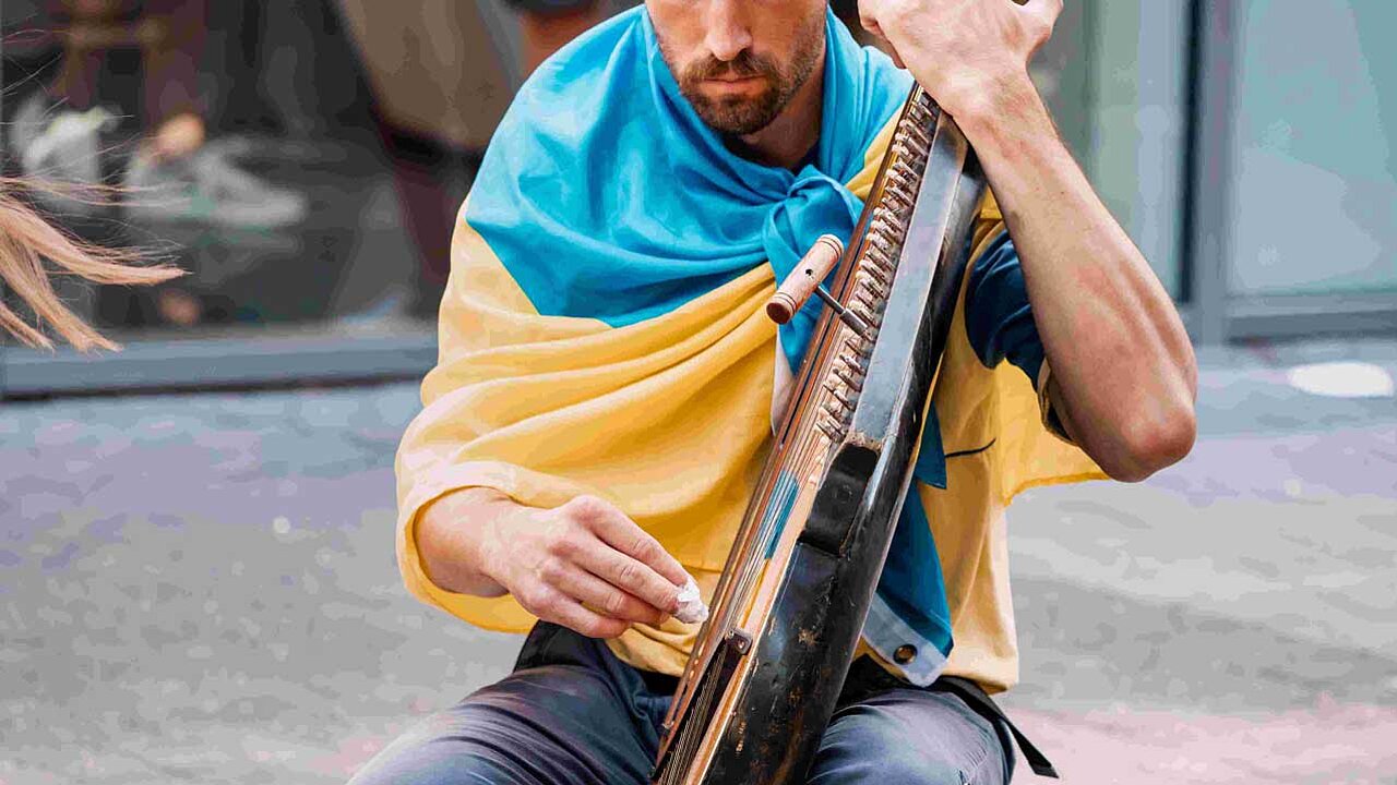 Eine Person sitzt in einer Fußgängerzone und spielt ein traditionelles Saiten-Instrument. Sie hat eine Ukrainische Flagge um den Hals gebunden