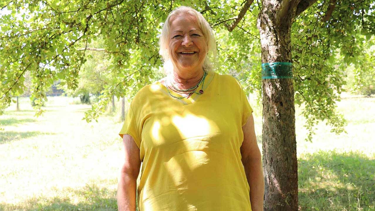 Eine Frau mit weißen Haaren und gelbem Oberteil steht unter einem Baum, viel Grün viel Licht, sie lächelt freundlich.