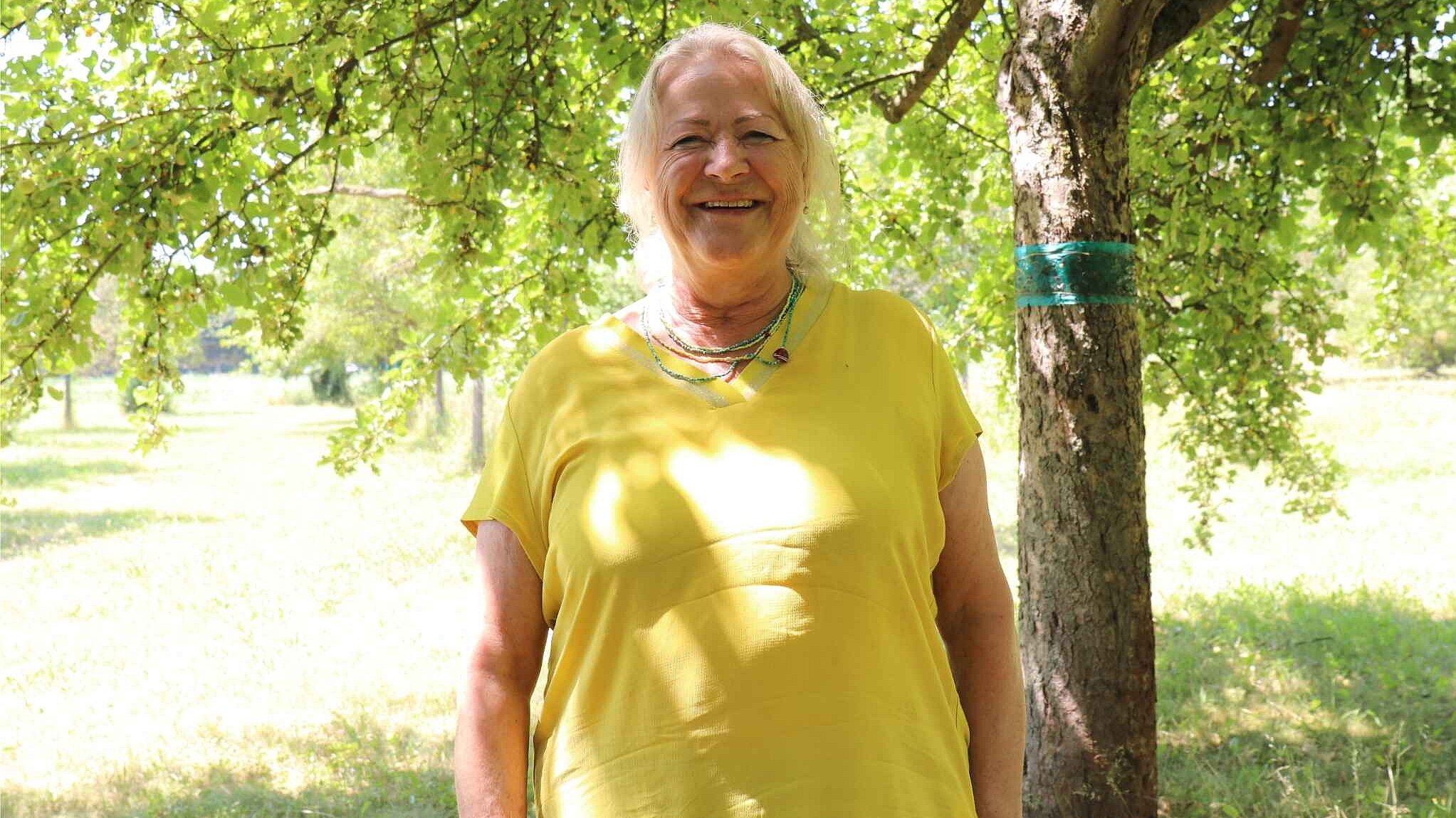 Eine Frau mit weißen Haaren und gelbem Oberteil steht unter einem Baum, viel Grün viel Licht, sie lächelt freundlich.