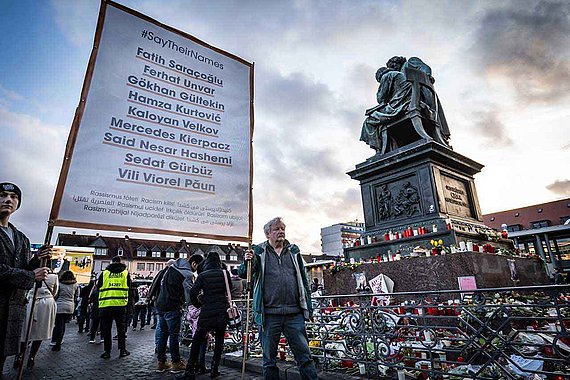 Plakat mit den Namen der Opfer aus Hanau, Blumen, Kränze und Kerzen am Brüder-Grimm-Denkmal auf dem Marktplatz 