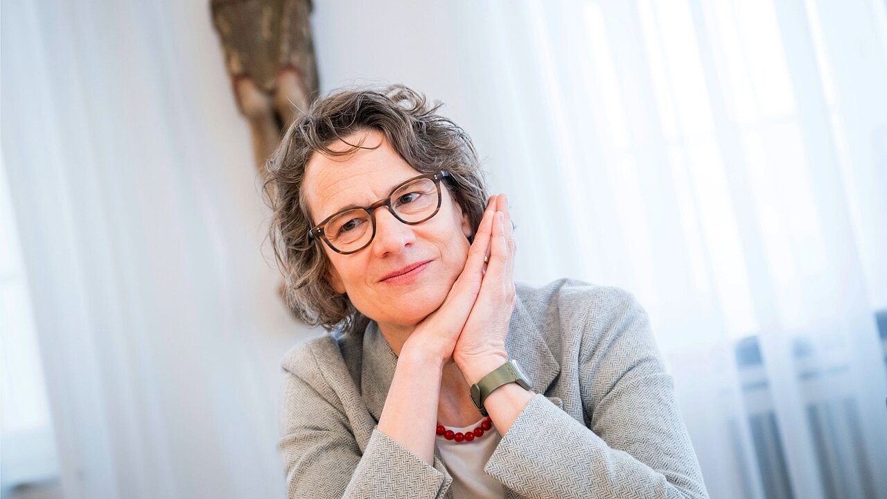 Eine Frau mit lockigen Haaren und Brille vor weißen Vorhängen