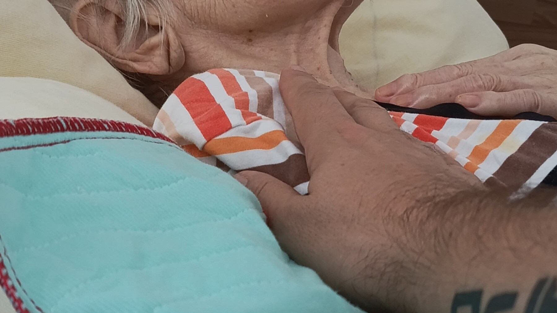 eine alte Person auf einem Bett. Nur Kinnpartie, Schultern und Brustkorb zu sehen. Eine Hand ruht auf der Brust. Eine andere Hand berührt sanft die Schulter der liegenden Person
