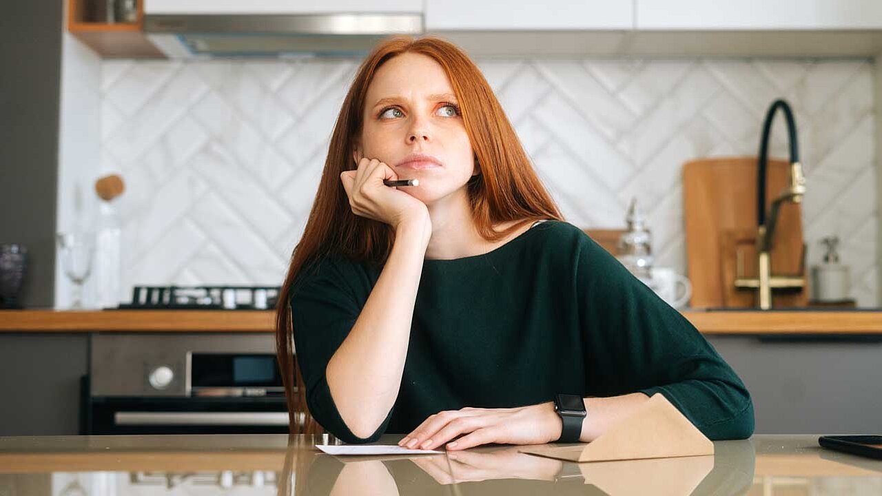 Vorderansicht einer rothaarigen Frau, die einen handgeschriebenen Brief schreibt, am Tisch mit Umschlag im Küchenraum mit modernem hellem Interieur 