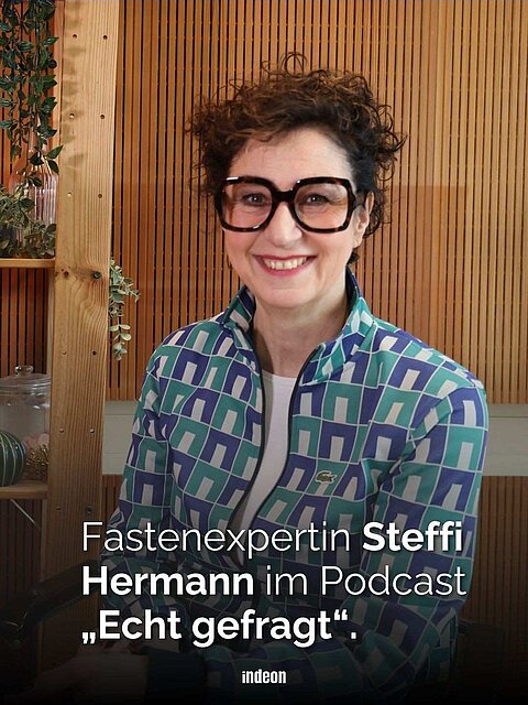 Eine Frau mit kurzen dunklen Locken, einer blaugrünen und einer großen schwarzen Brille vor einer Holzwand. Text: Fastenexpertin Steffi Hermann im Podcast „Echt gefragt“.