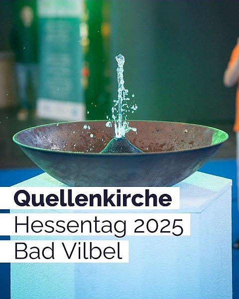 Eine sprudelnde Wasserschale auf einem Sockel. Beschienen von grünem und blauem Licht. Text: "Quellenkirche Hessentag 2025 Bad Vilbel."