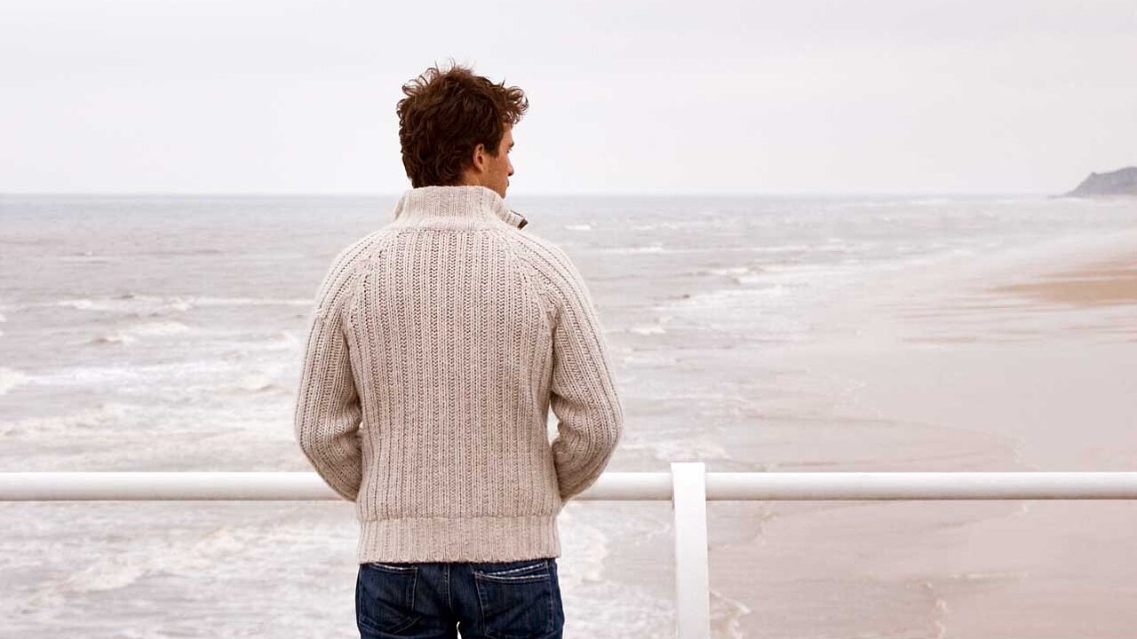 An der Strandpromenade steht ein Mann mit Blick aufs Meer. Er hat die Hände in den Taschen.