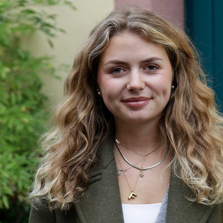 Portrait von Floriane. Sie ist blond, die Haare reichen über die Schultern. Sie trägt mehrere Ketten.