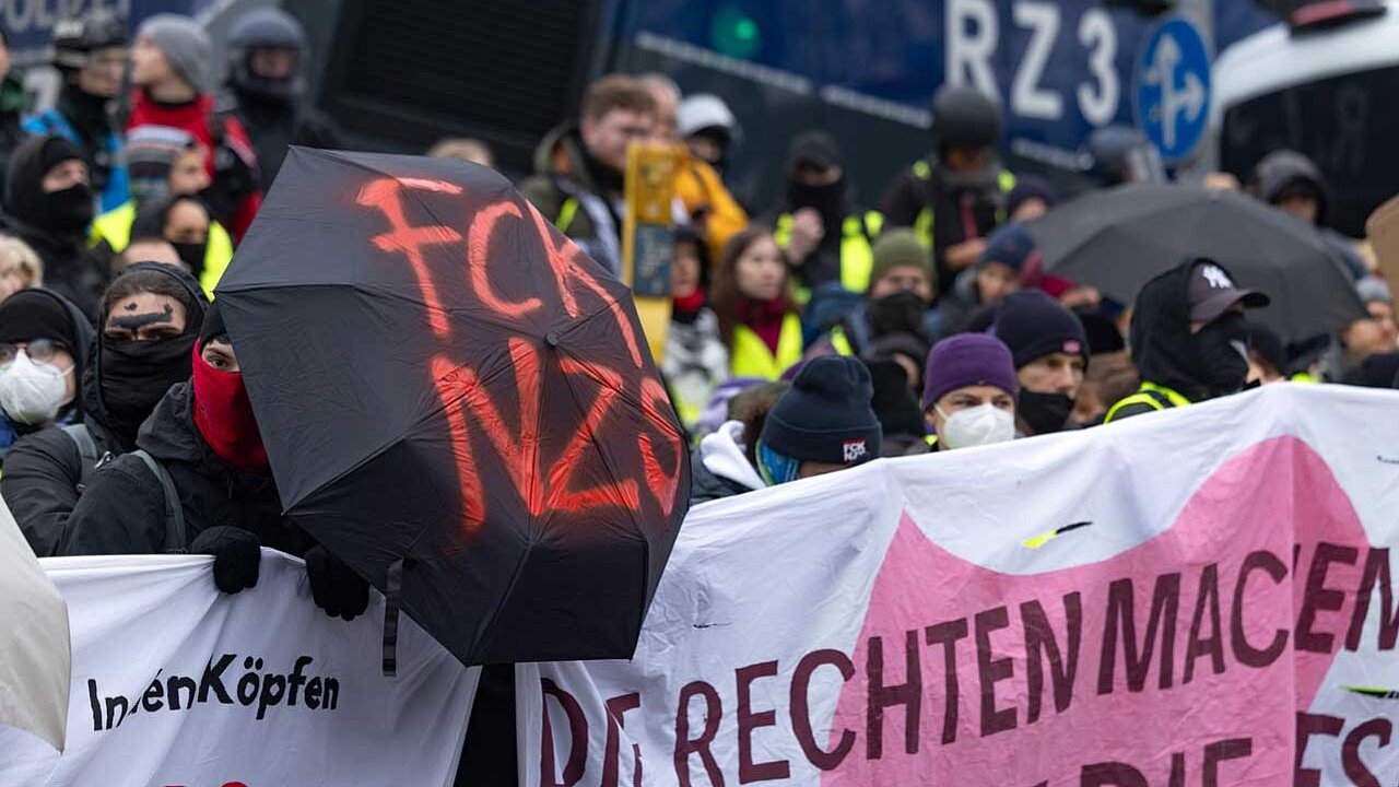 Protestkundgebung mit Schildern und Fahnen, darunter ein schwarzer Regenschirm mit roter Aufschrift 'FCK NZS', im Hintergrund ein Polizeifahrzeug.