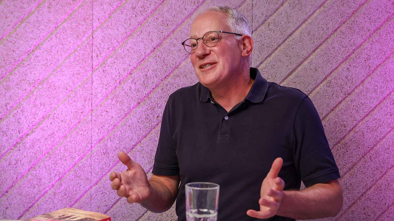 Ein Mann mit schwarzem Shirt, weißen Haaren und Brille vor pinker Wand.