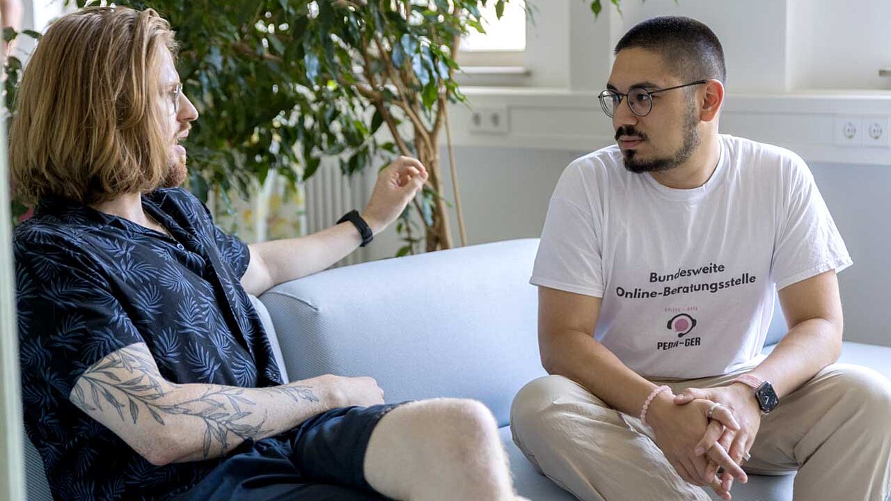 Kiril und Furkan sitzen auf einer hellblauen Couch und sprechen miteinander.