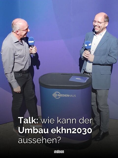Zwei ältere Männer vor einer lila Wand mit Mikrophonen in der Hand. Zwischen ihnen ein Pult. Text: Talk: wie kann der Umbau ekhn2030 aussehen