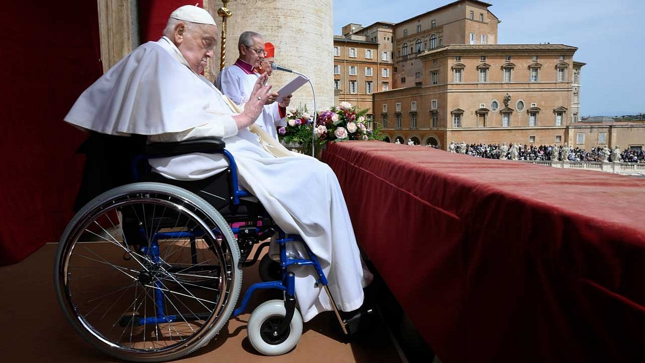 Papst Franziskus sitzt im Rollstuhl und spricht den Segen in ein Mikrofon. 