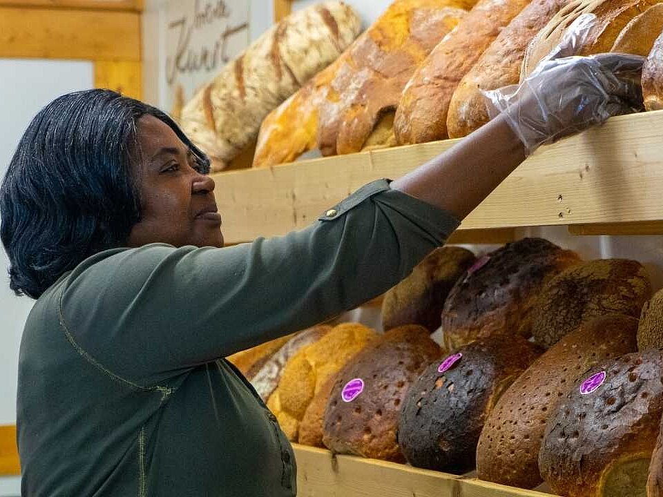 Eine Mitarbeiterin mit schwarzen schulterlangen Haaren räumt Brot in die Backtheke ein.