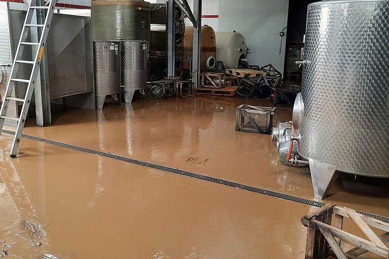 Tanks und Stahlbehälter beim Winzerverein, alles dreckig. Der Boden ist überzogen von einer dicken Schlammschicht mit Fußspuren darin. Mehrere Kisten liegen teilweise kaputt auf dem Boden herum. Ein Bild der Zerstörung