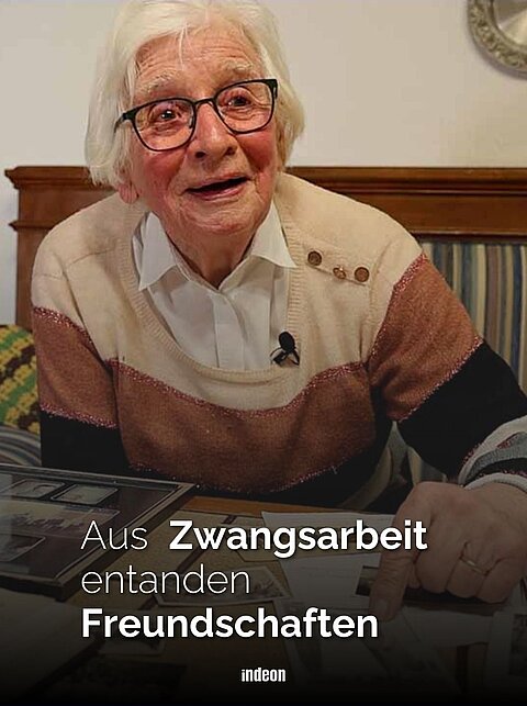 Auf dem Bild ist Ruth Kreiselmaier zusehen. Die ältere Dame hat weißes Haar und schaut freundlich in die Kamera. Sie sitzt an einem Tisch und zeigt auf alte Dokumente, die vor ihr auf der Tischplatte ausgelegt sind. 