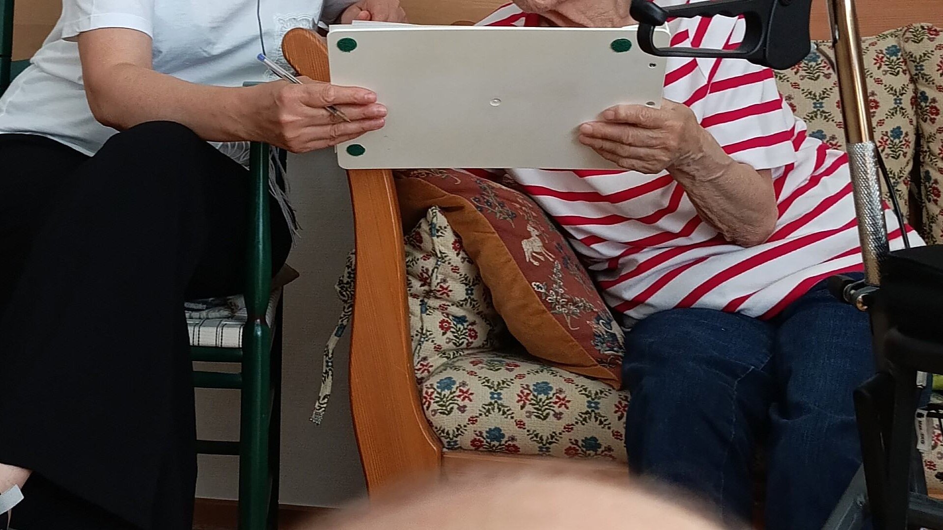 Zwei Personen sitzen nebeneinander, von den Schultern abwärts zu sehen. Sie zeigen einander etwas auf einer Tafel