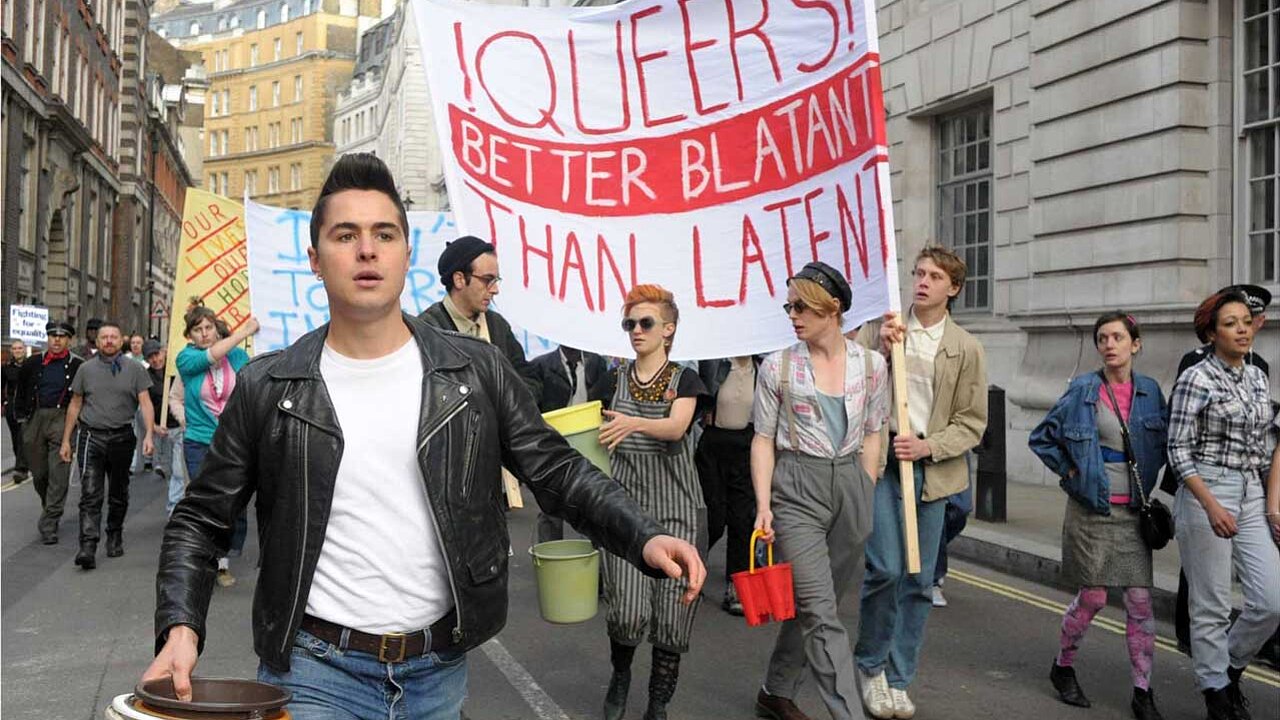Eine Gruppe von Demonstrierenden: im Vordergrund eine Person mit schwarzer Lederjacke, weißem Shirt und blauer Jeans, sie trägt einige Eimer in der Hand. Dahinter eine größere Gruppe mit einem Banner: „Queers! Better blatant than Latent