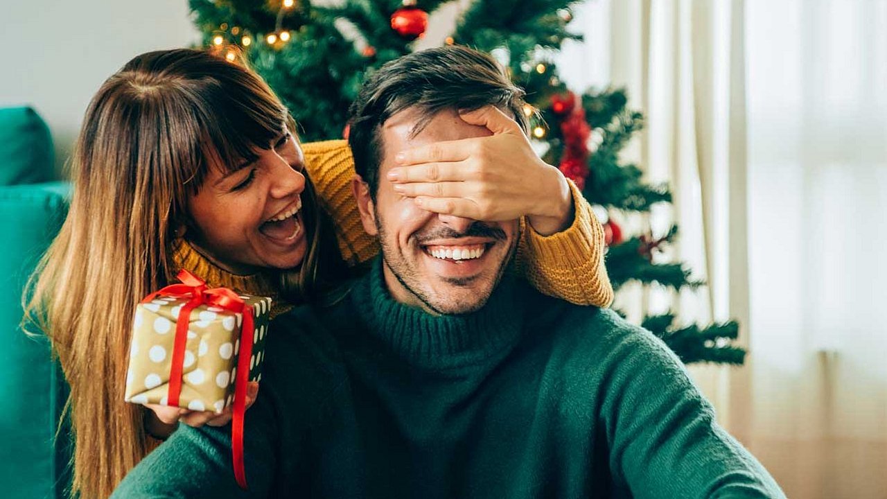 Die junge Frau steht hinter ihrem Freund mit einem Geschenk in der einen Hand und bedeckt seine Augen mit der anderen Hand.