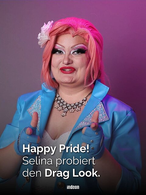 Redakteurin Selina mit pinker Perücke, blau-bunter Lederjacke, prächtigem Schmuck und knalligem Makeup lacht in die Kamera. Text: Happy Pride, Selina probiert den Drag Look