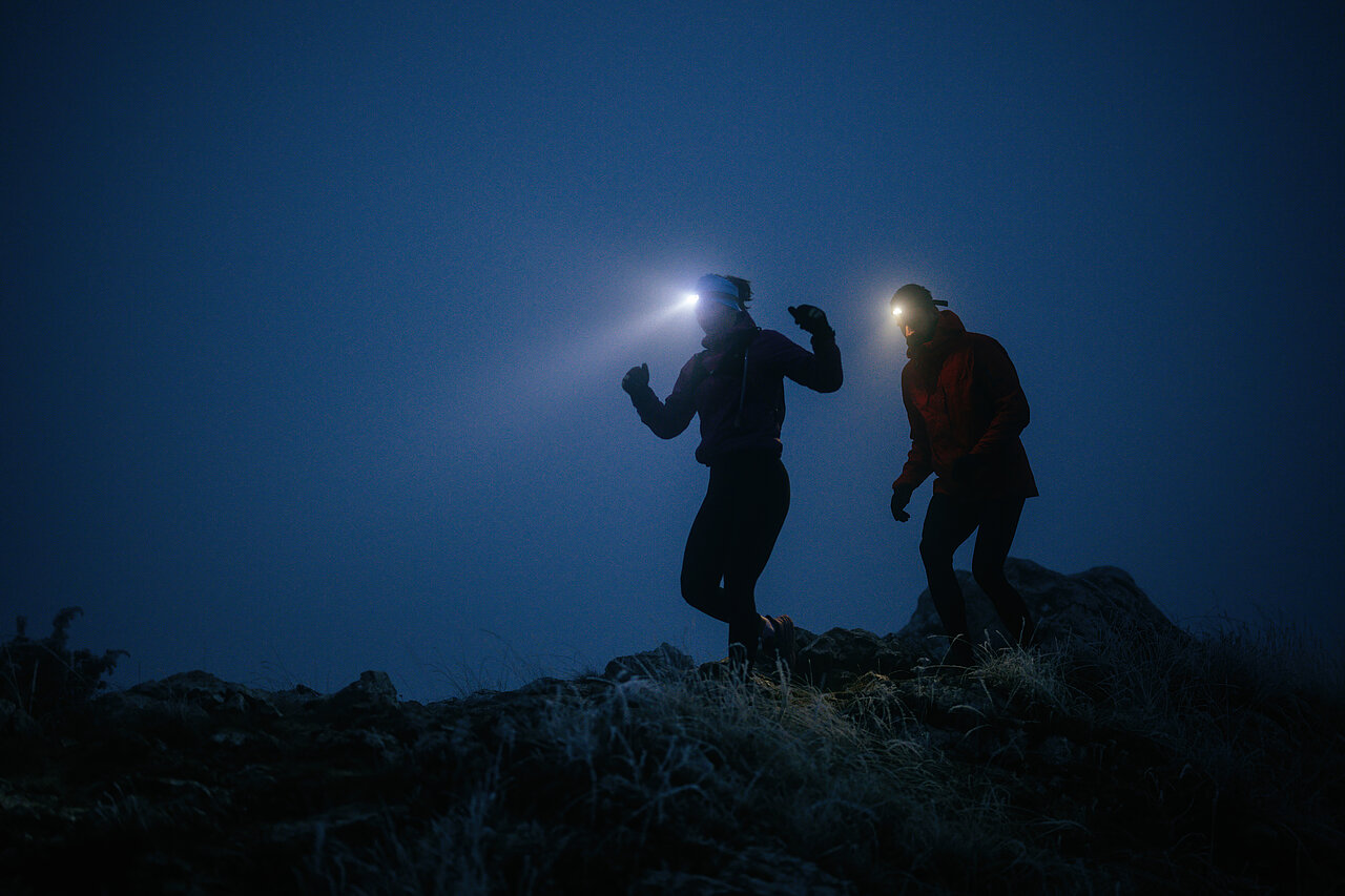 Zwei Personen wandern nachts über einen felsigen Weg und tragen Stirnlampen, deren Licht in die Dunkelheit strahlt.