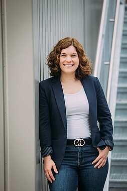 Maren Busch steht mit weißer Bluse und Blauen Jackett an eine Wand gelehnt