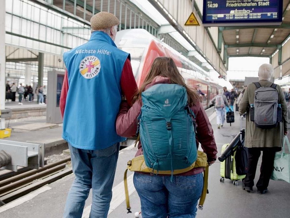 Ein Mann in einer blauen Weste, auf der das Logo der Bahnhofmission zu  sehen ist, begleitet eine blinde junge Frau, die einen türkisfarbenen Rucksack trägt zum Bahnsteig. Die blinde Frau ist bei dem Ehrenamtlichen eingehakt. Sie sind von hinten zu sehen, wie sie zum richtigen Gleis gehen.