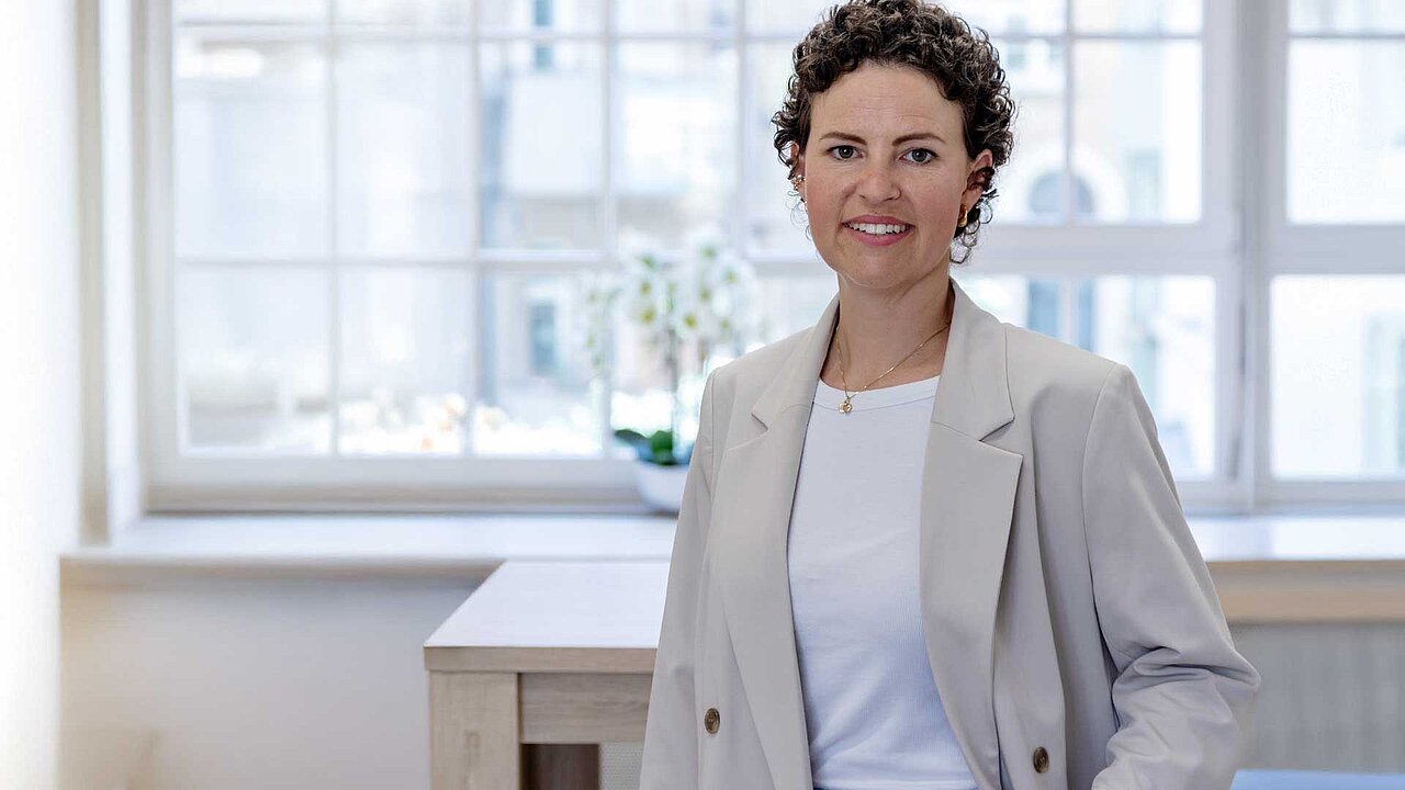 Sarah steht vor einem Fenster und lächelt in die Kamera