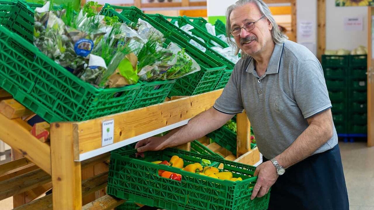 Ein Mitarbeiter der Tafel, ein älterer Mann im hellgrauen T-Shirt räumt grüne Gemüsekisten ein.