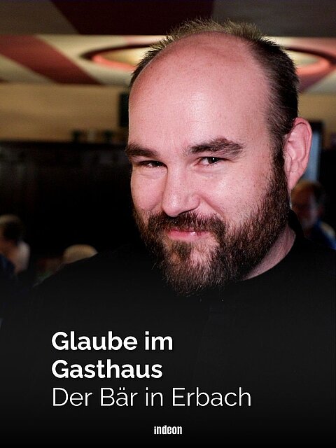 Ein Mann mit Halbglatze, dunklem Bart, dunklem Shirt und freundlichem Lächeln steht in einem Gastraum. Text: Glaube im Gasthaus: der Bär in Erbach