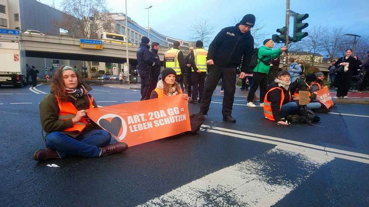 Zwei frauen sitzen mit einem Plakat auf der Straße.Ein Polizist spricht sie an.