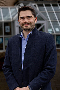 Portraitfoto von Lukas Daum mit blauem Hemd und blauem Mantel vor dem Rathaus in Gilserberg