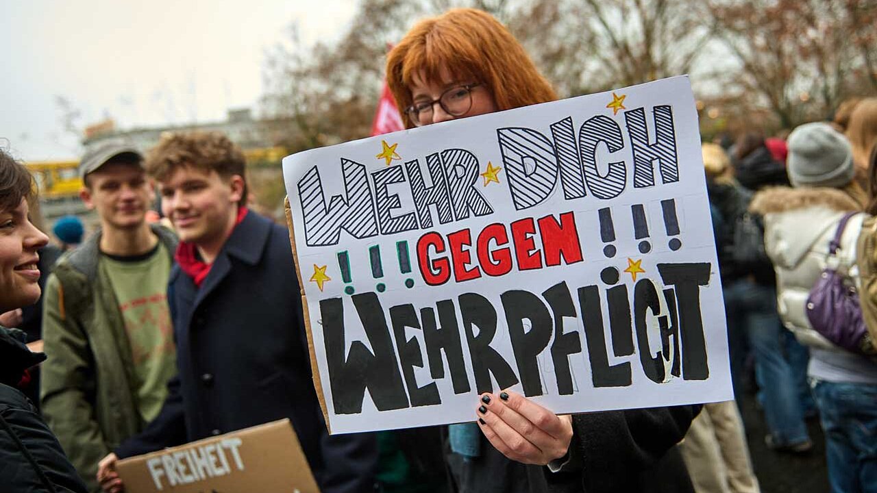 Person mit roten Haaren hält ein Schild in die Kamera mit dem Text: „Wehr dich gegen Wehrpflicht!“