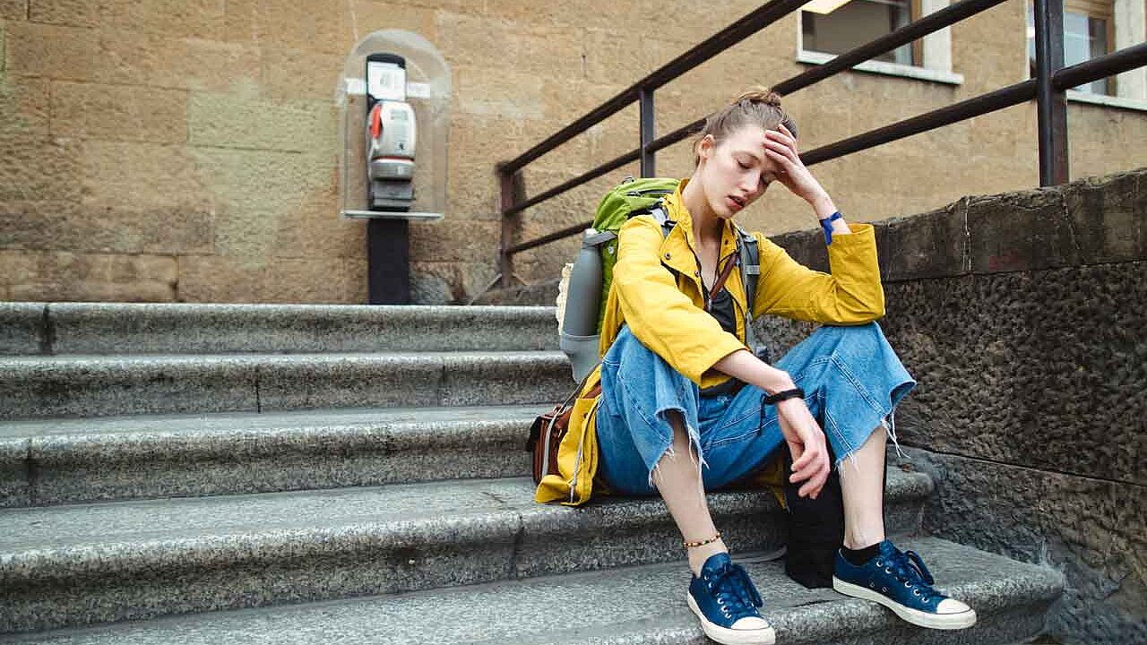 Das Bild zeigt eine junge Person, die mit Rucksack auf einer Steintreppe sitzt und sich erschöpft oder nachdenklich die Hand an die Stirn hält.